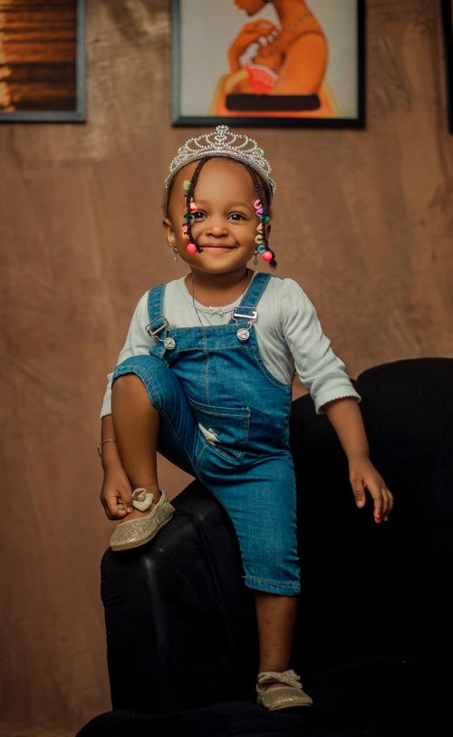 Free Adorable child wearing a tiara and denim overalls posing on a black chair indoors. Stock Photo