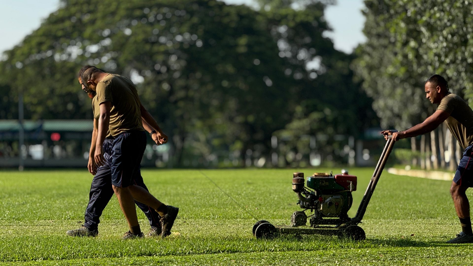 Two men mowing lawn in a sunny park setting in Dhaka, Bangladesh.