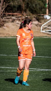 A female soccer player in orange uniform plays on a sunlit outdoor field.