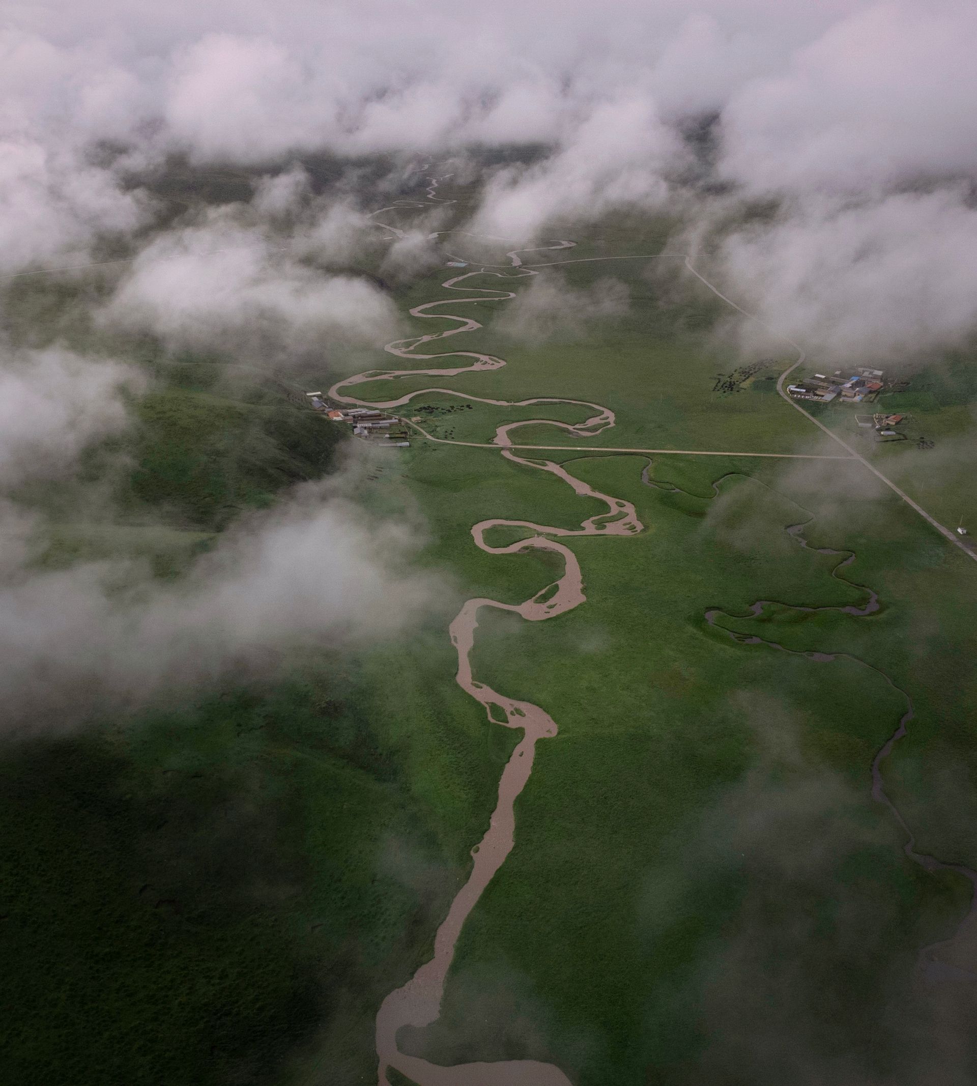 Aerial view of a winding river cutting through lush green grasslands beneath misty clouds