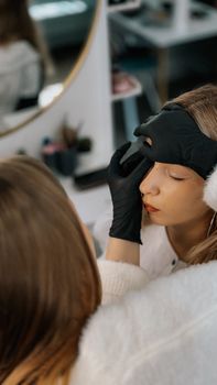 Young beautician wearing gloves applies eye makeup to a teen girl in a beauty salon.