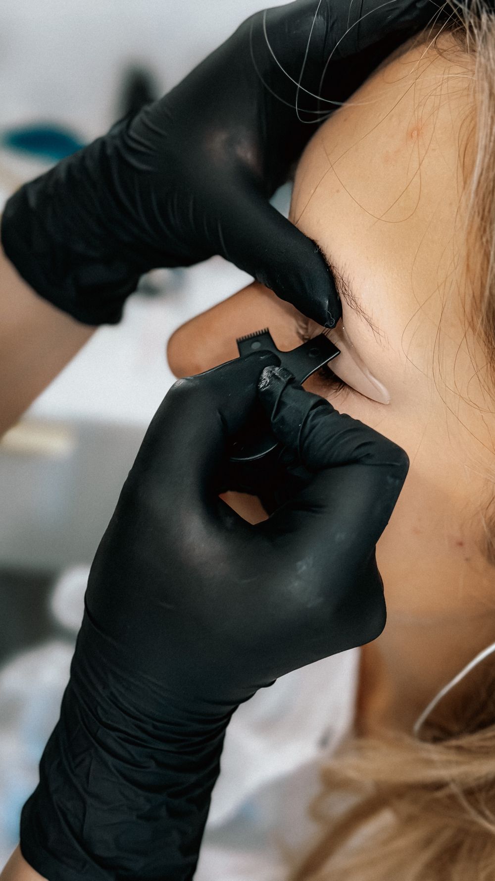 Close-up of a black-gloved practitioner using a small brow tool near a client's eyebrow and temple area.