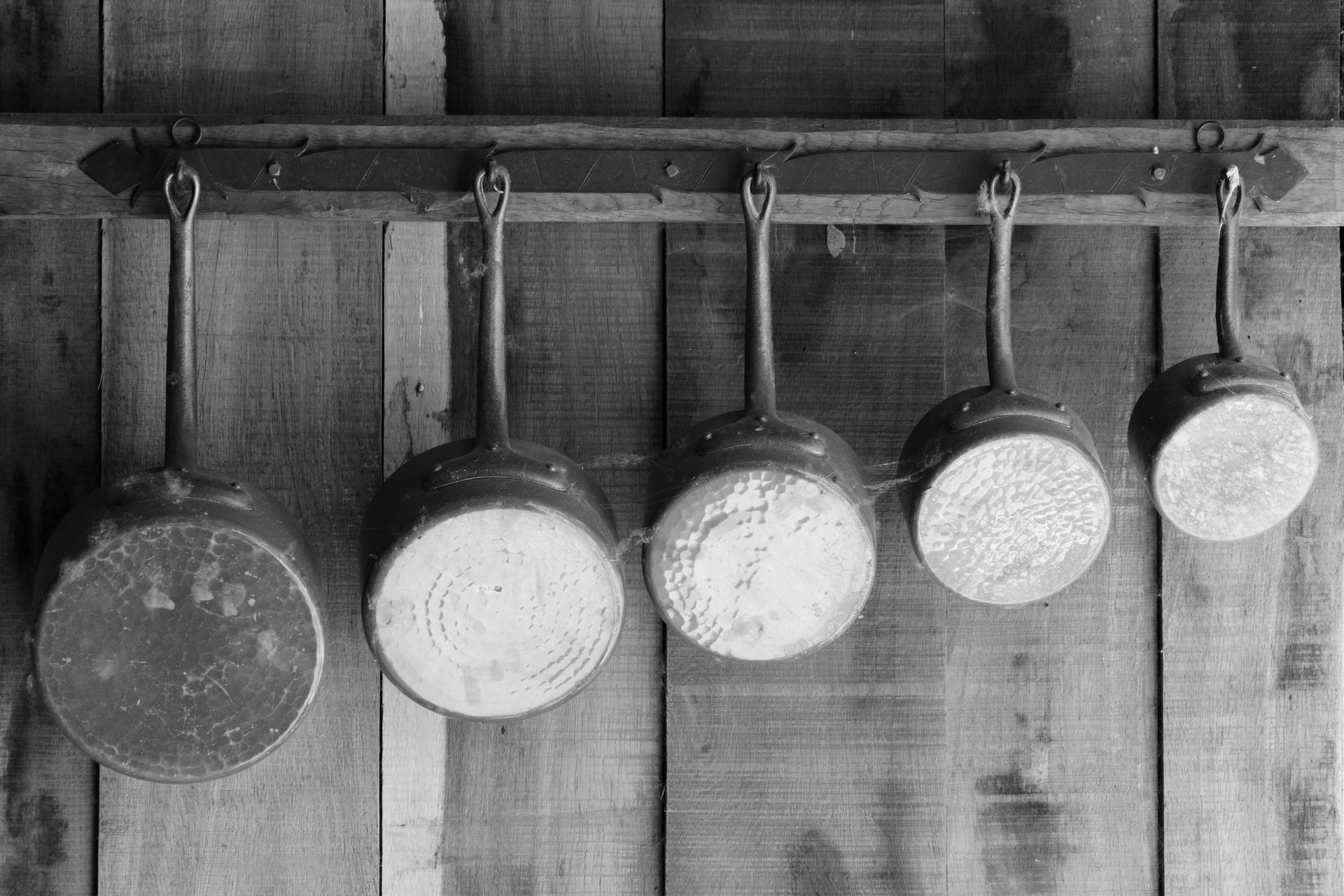 Monochrome photo of vintage copper pots hanging on a rustic wooden wall, showcasing old-world charm.