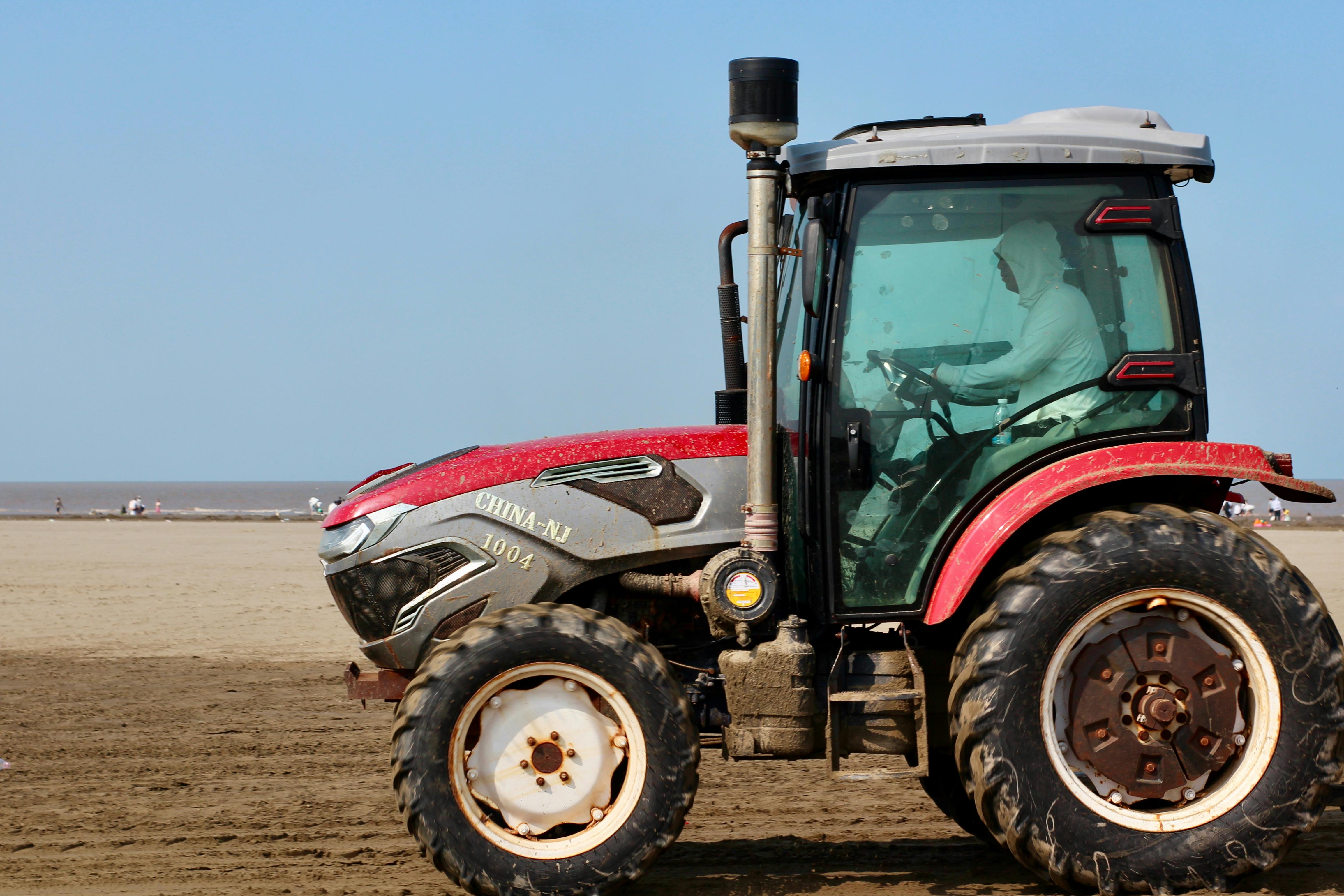 A lone red tractor drives along a sandy beach under a bright blue sky.