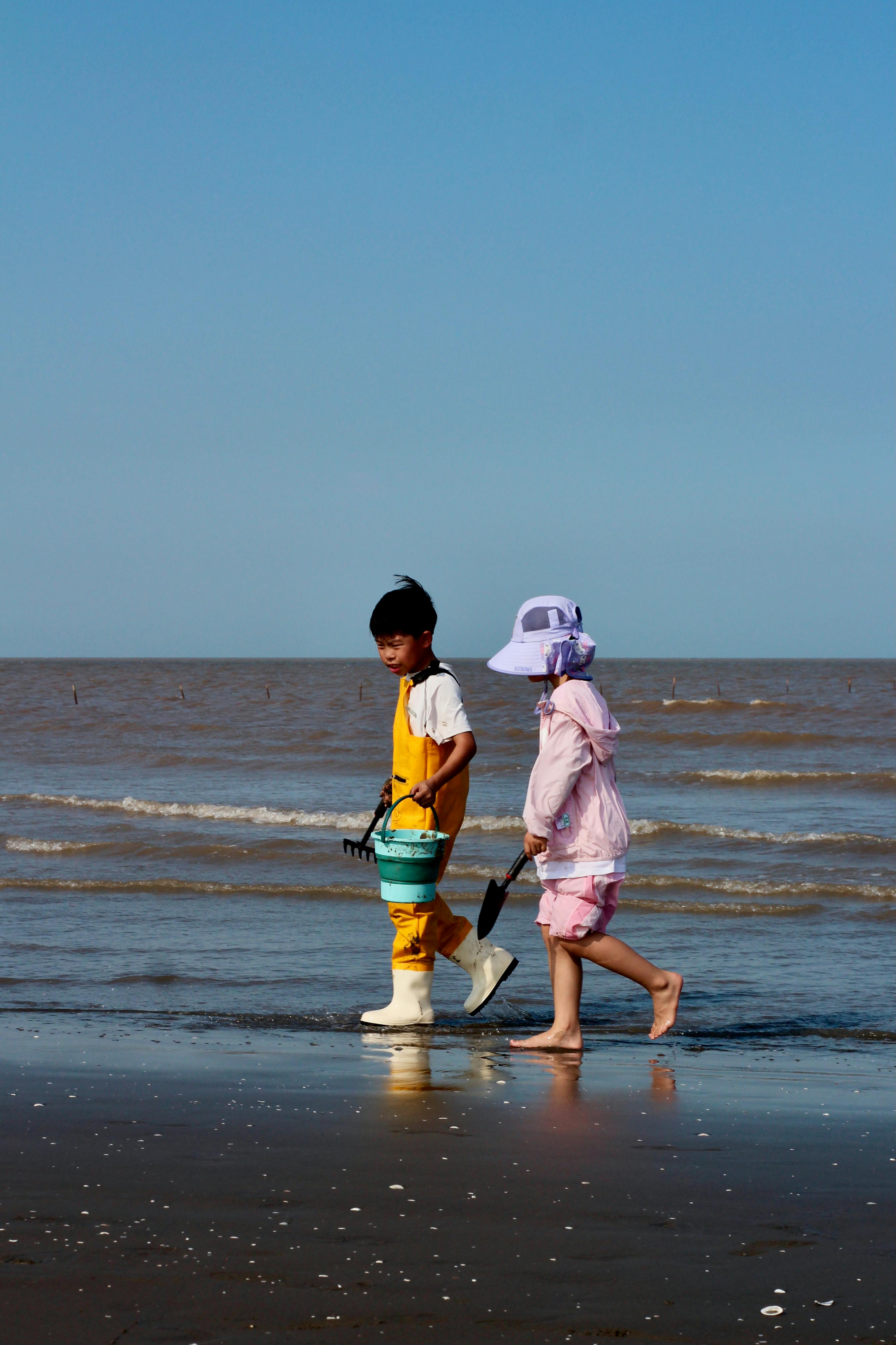 ฟรี เด็กสองคนสำรวจชายหาดทรายพร้อมถังน้ำใต้ท้องฟ้าสีฟ้าใส คลังภาพถ่าย