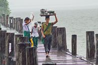 People crossing U Bein Bridge in Mandalay during a rainy day, carrying goods.