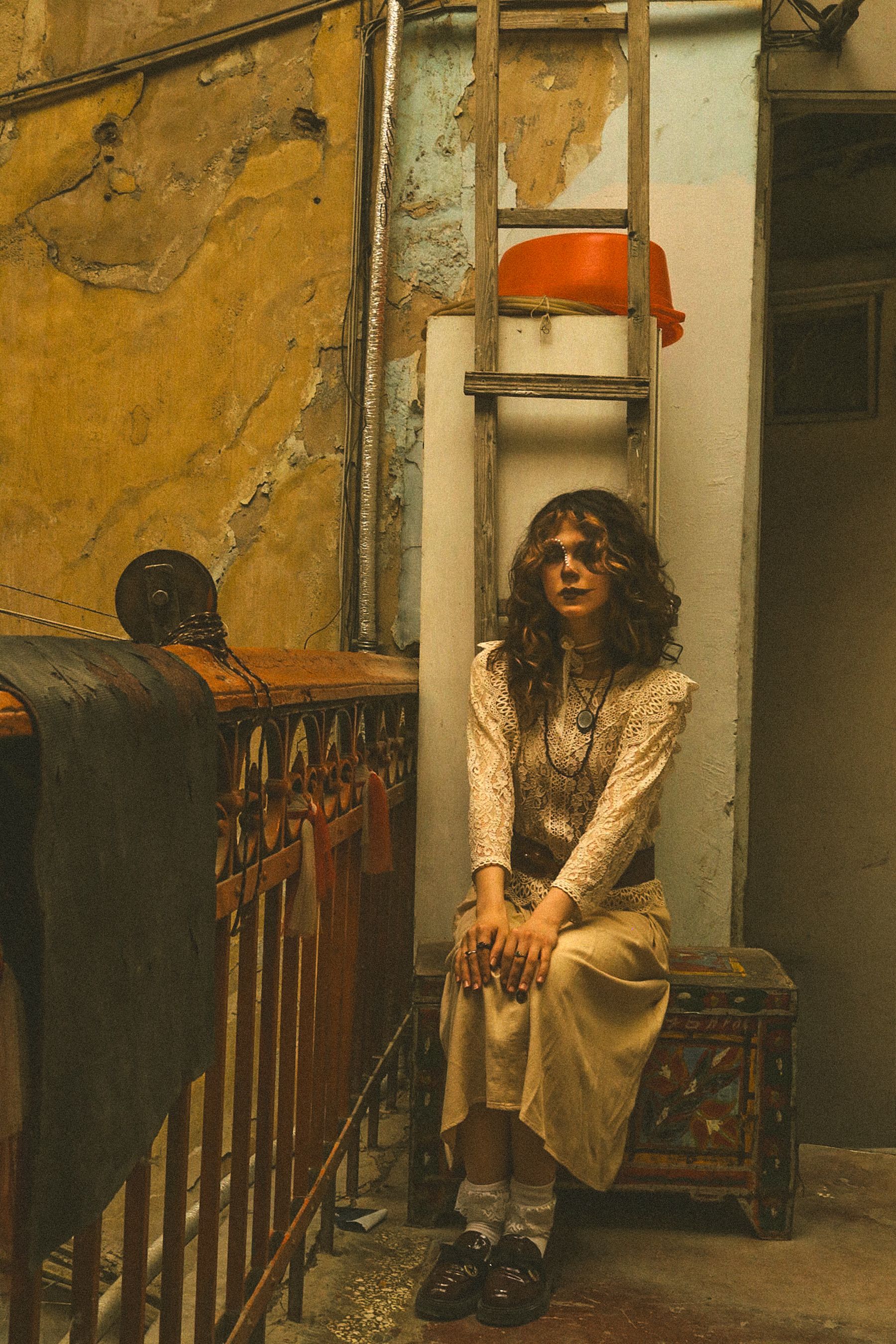 A woman in vintage attire seated indoors with a rustic backdrop.