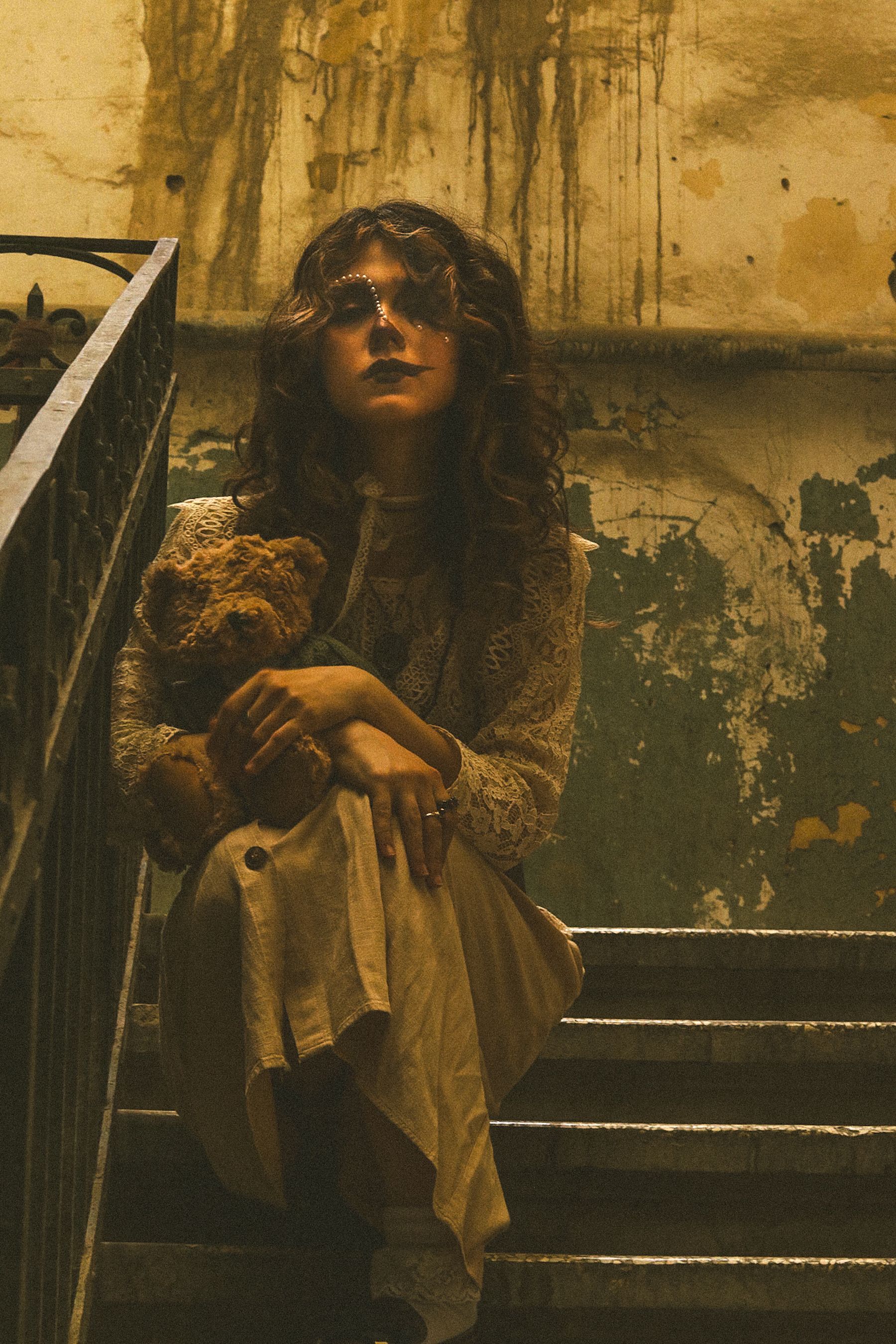 A woman in vintage attire sits on stairs with a teddy bear in a dramatic, moody setting.