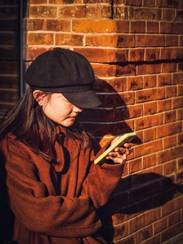 Woman leaning against a brick wall, holding a smartphone, wearing a brown coat and black hat.
