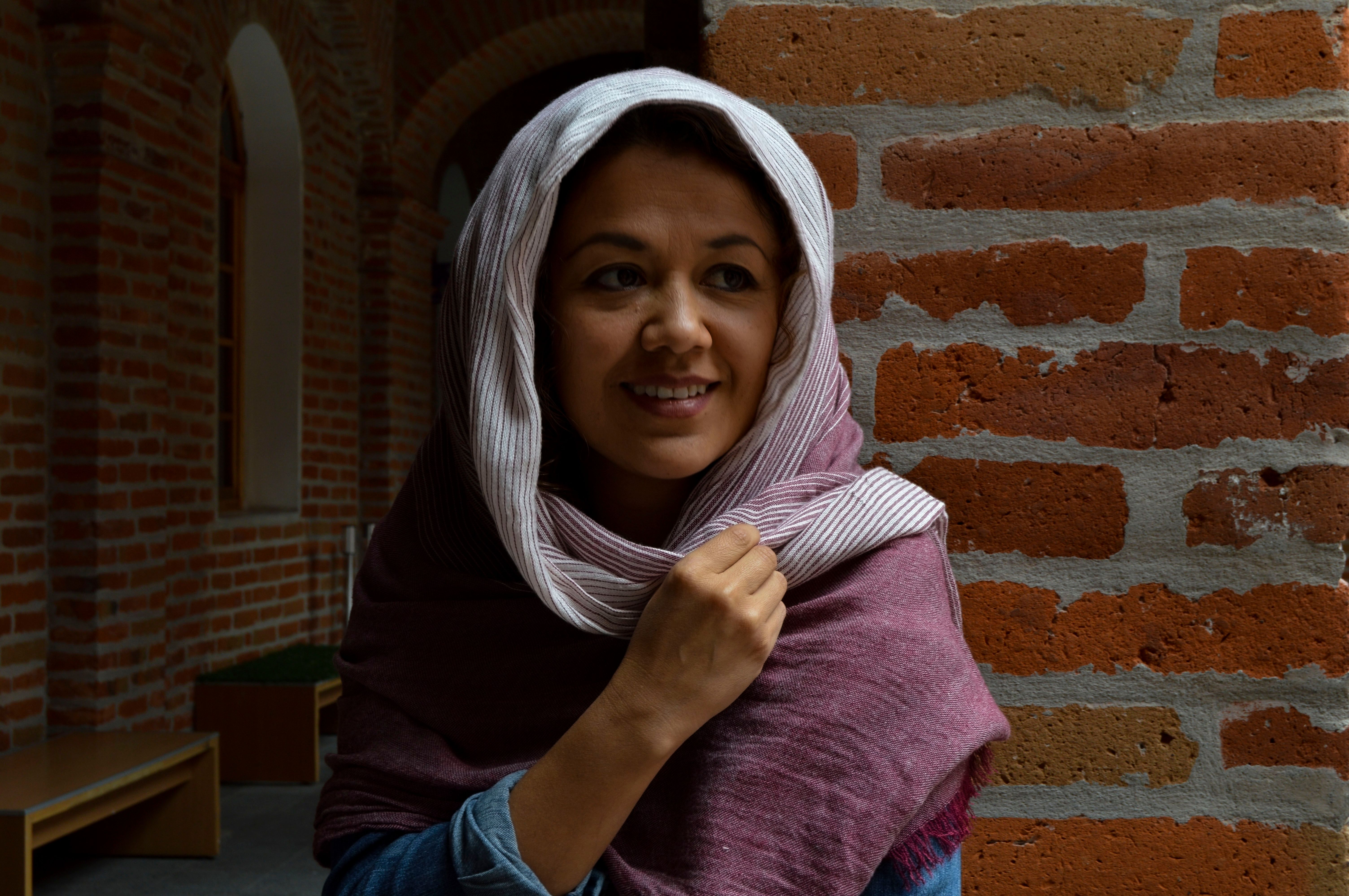 Portrait of a young woman in a scarf against a rustic brick wall indoors.
