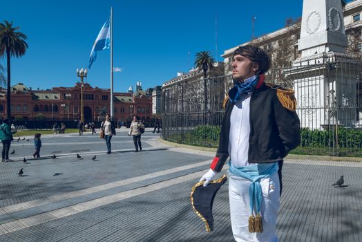 Man in historical costume at Plaza de Mayo, Buenos Aires with landmarks and Argentine flag.