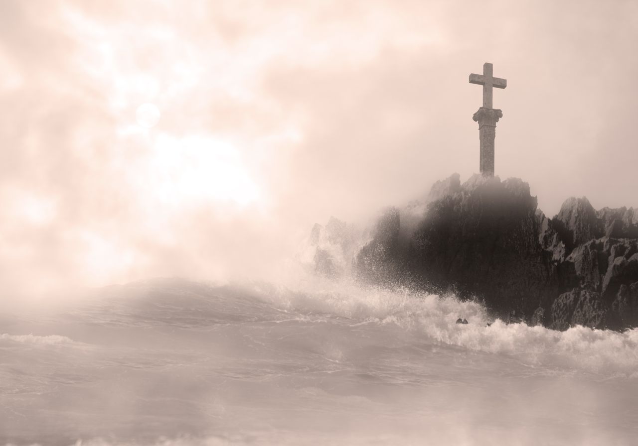 moody image of a cross on rocks amidst crashing waves and misty seas