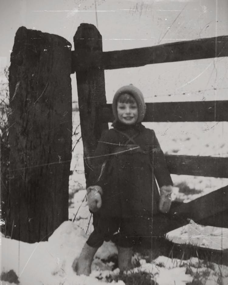 Grayscale Photography Of A Child Standing On Snow Beside A Wooden Fence