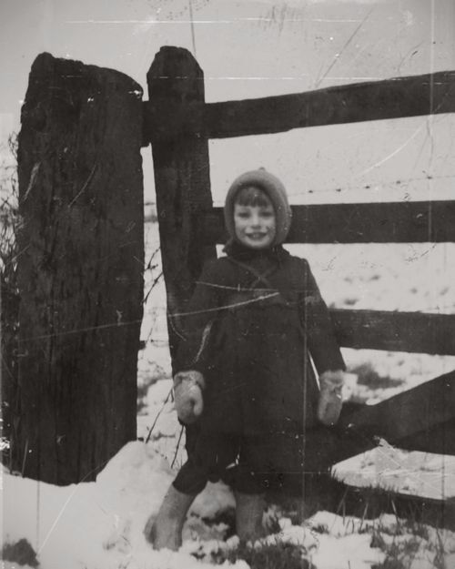 Free Classic black and white photo of a child in winter attire standing by a wooden fence in the snow. Stock Photo