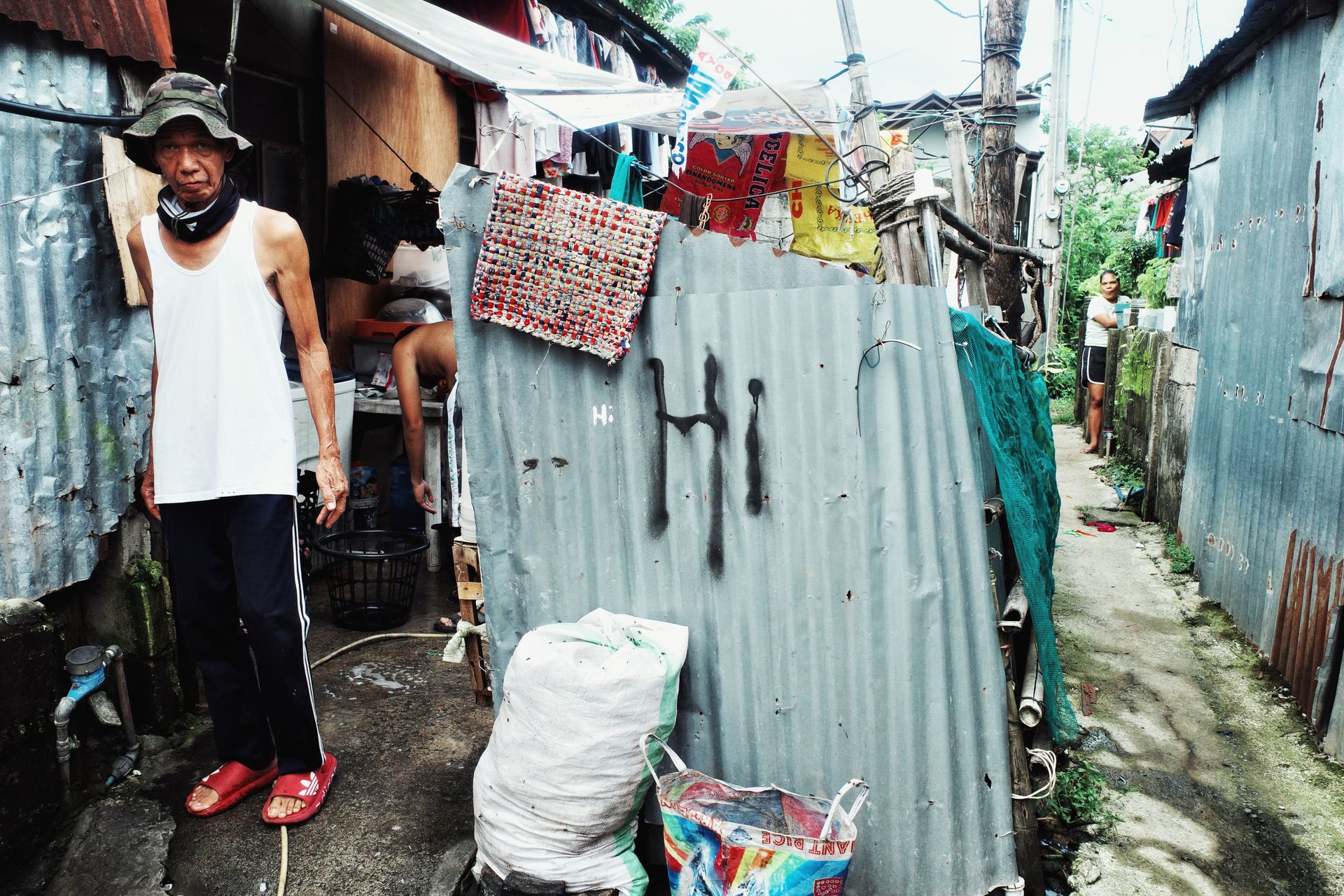 Residents standing in an urban alleyway between makeshift homes, surrounded by corrugated metal structures.