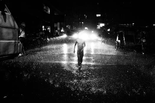 A lone figure walks through a rainy street illuminated by headlights at night.