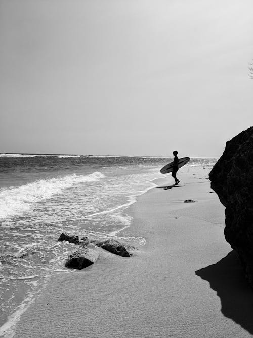 サーフボードを持って海岸に立っている人のグレースケール写真