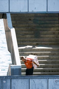 A worker in an orange uniform carries boxes up a sunny outdoor staircase by the water.