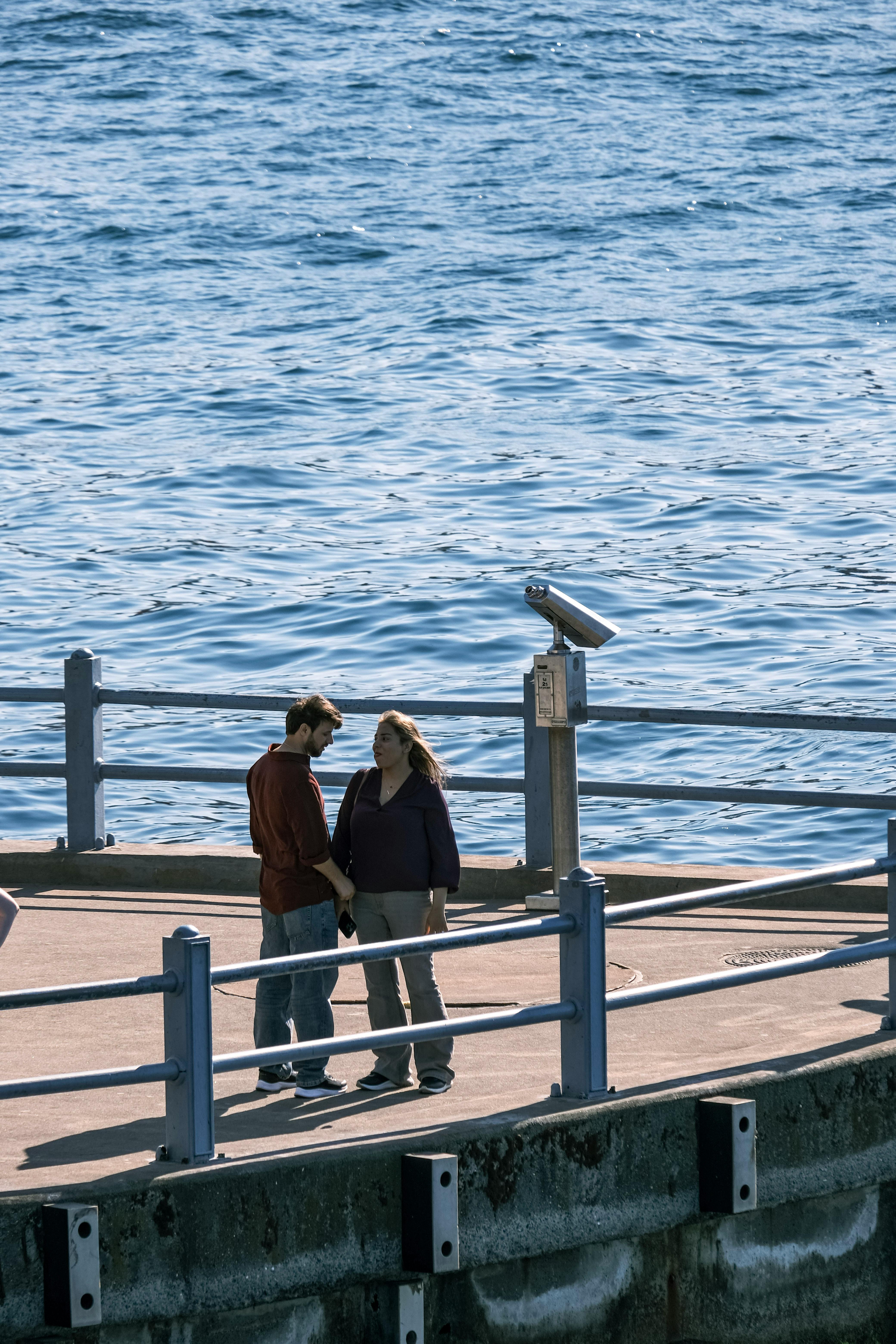 A couple enjoys a quiet moment on a sunny waterfront pier, holding hands and engaging in conversation.