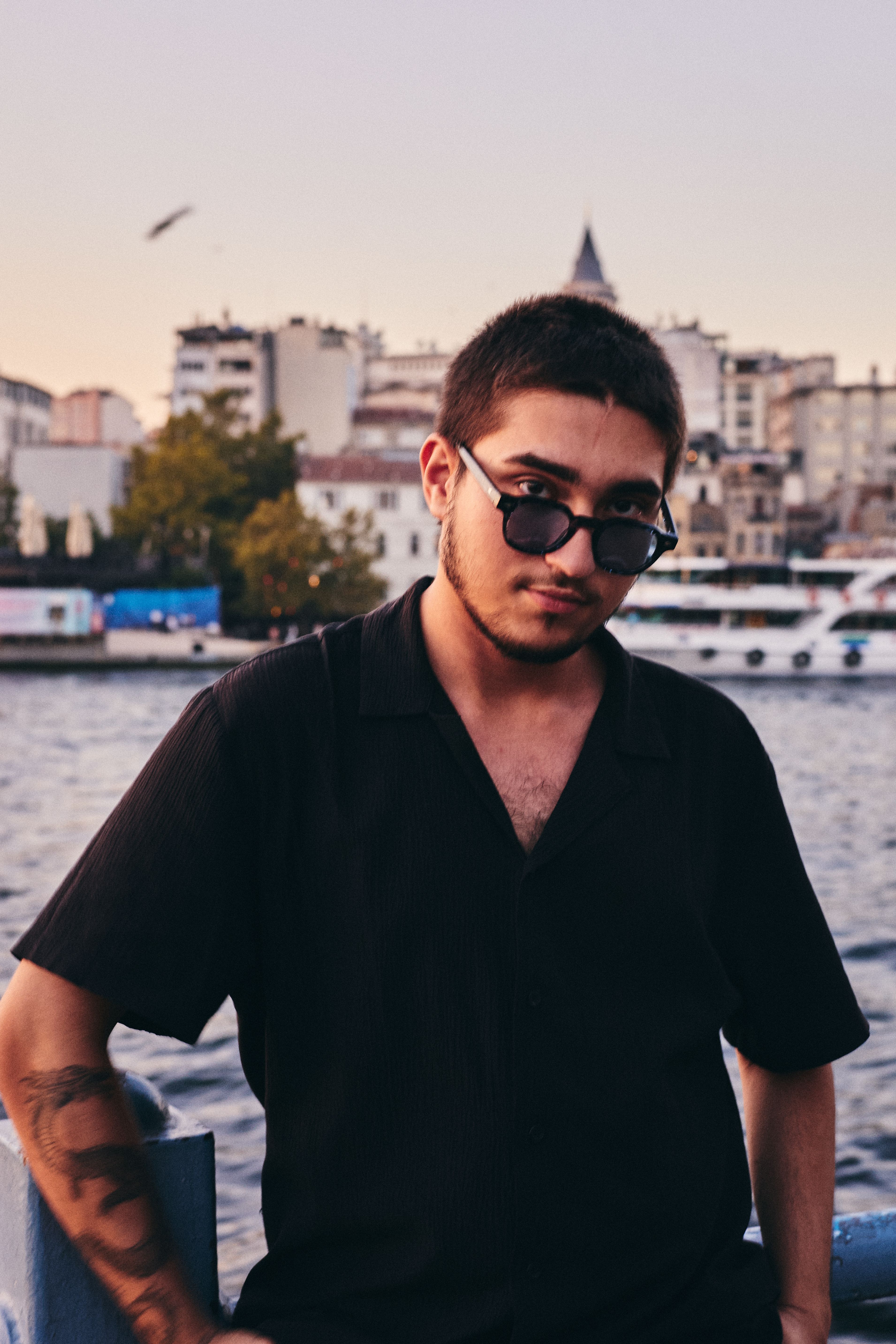 Stylish young man in sunglasses posing by the Bosphorus in İstanbul at dusk with cityscape background.