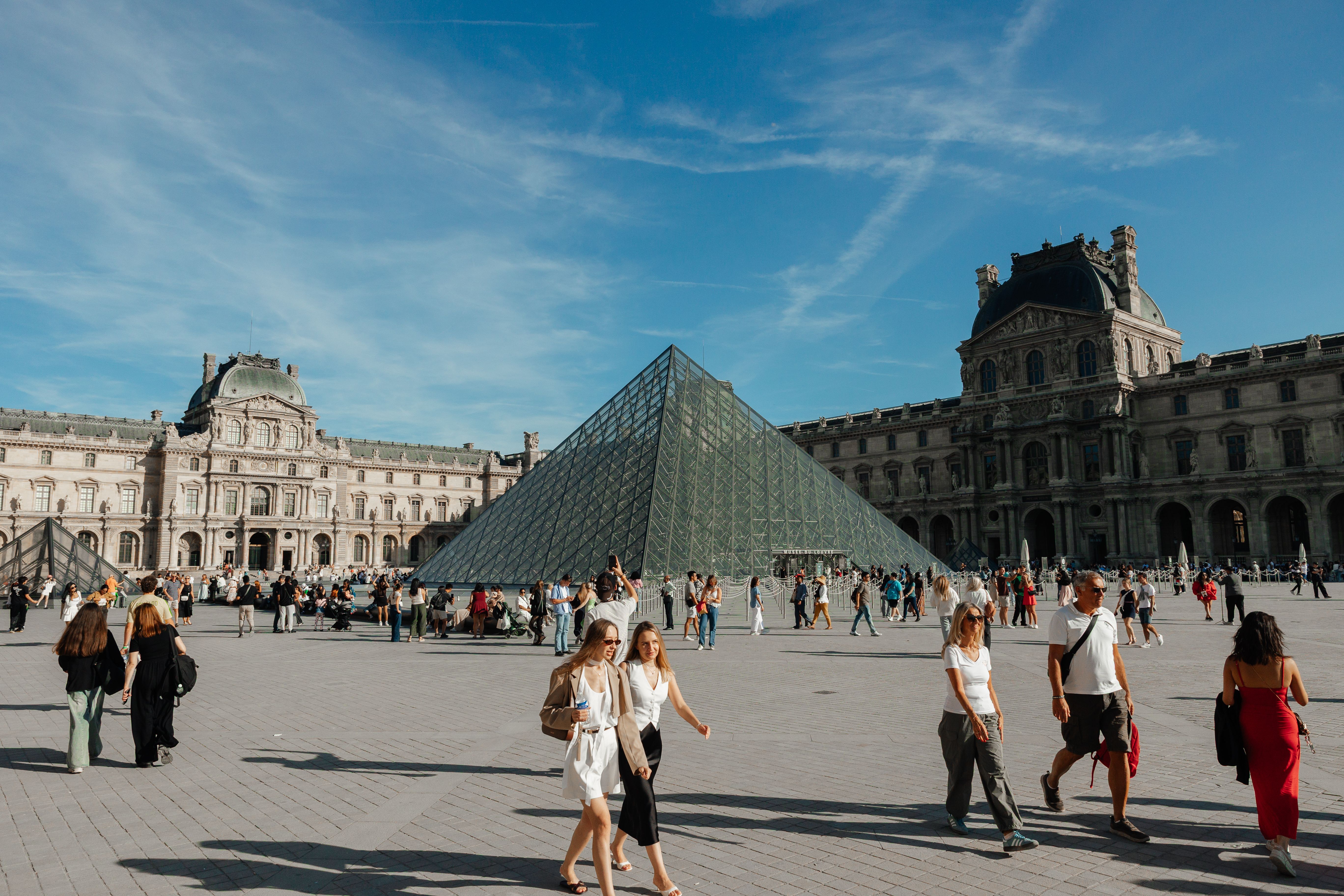 Crowds gather around the iconic glass Pyramid of the Louvre Museum in Paris, France on a sunny day.