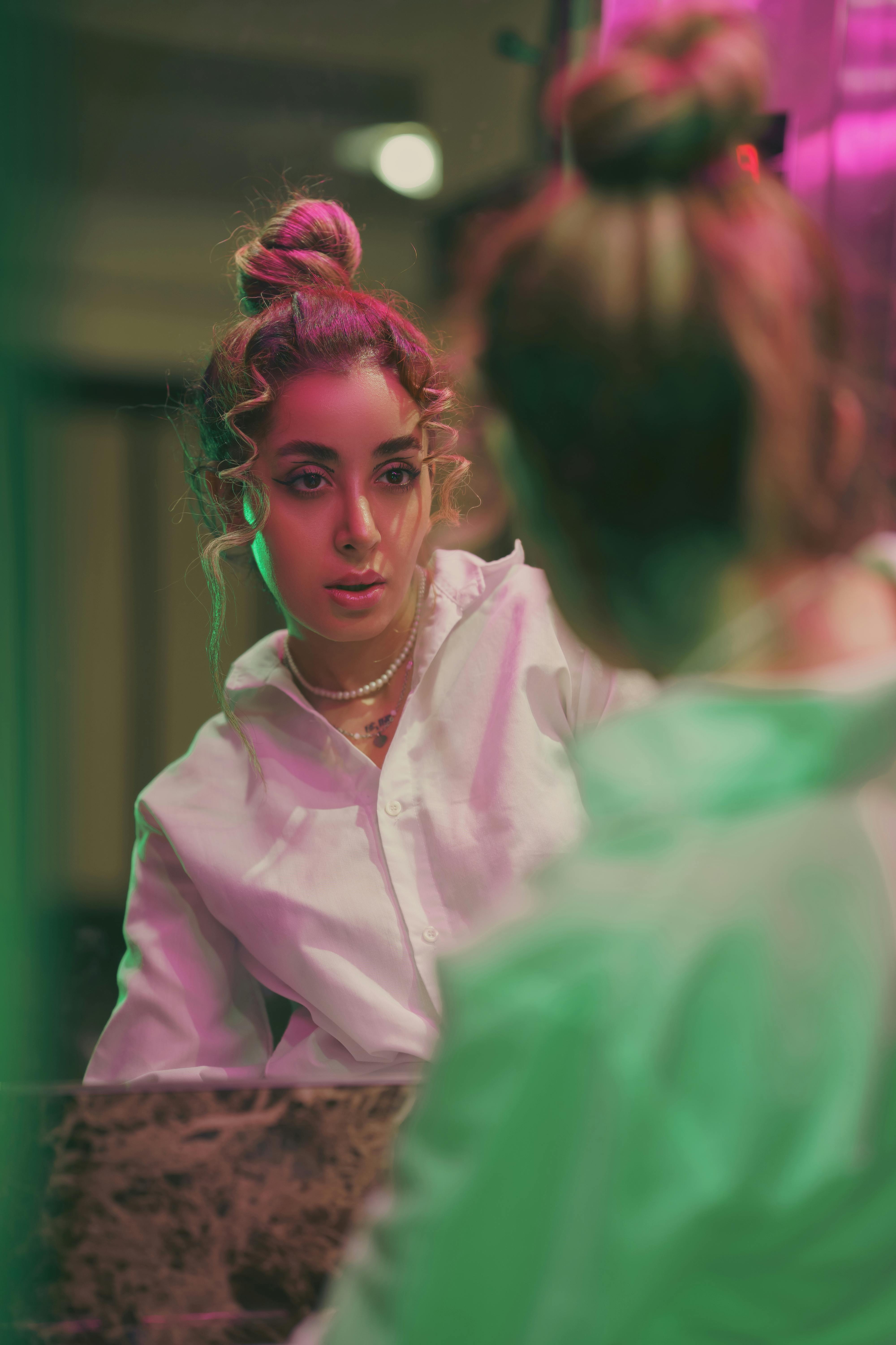 A stylish woman in a white shirt looks at her reflection in a neon-lit mirror, exuding urban fashion vibes.