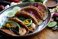 Colorful Mexican tacos with shrimp and avocado served on a metal plate, garnished with lime and flowers.