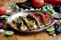 Three flavorful Mexican tacos on a plate surrounded by fresh ingredients.