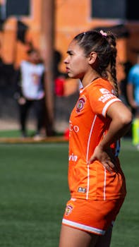 A young female soccer player in an orange uniform stands on the field during a match.