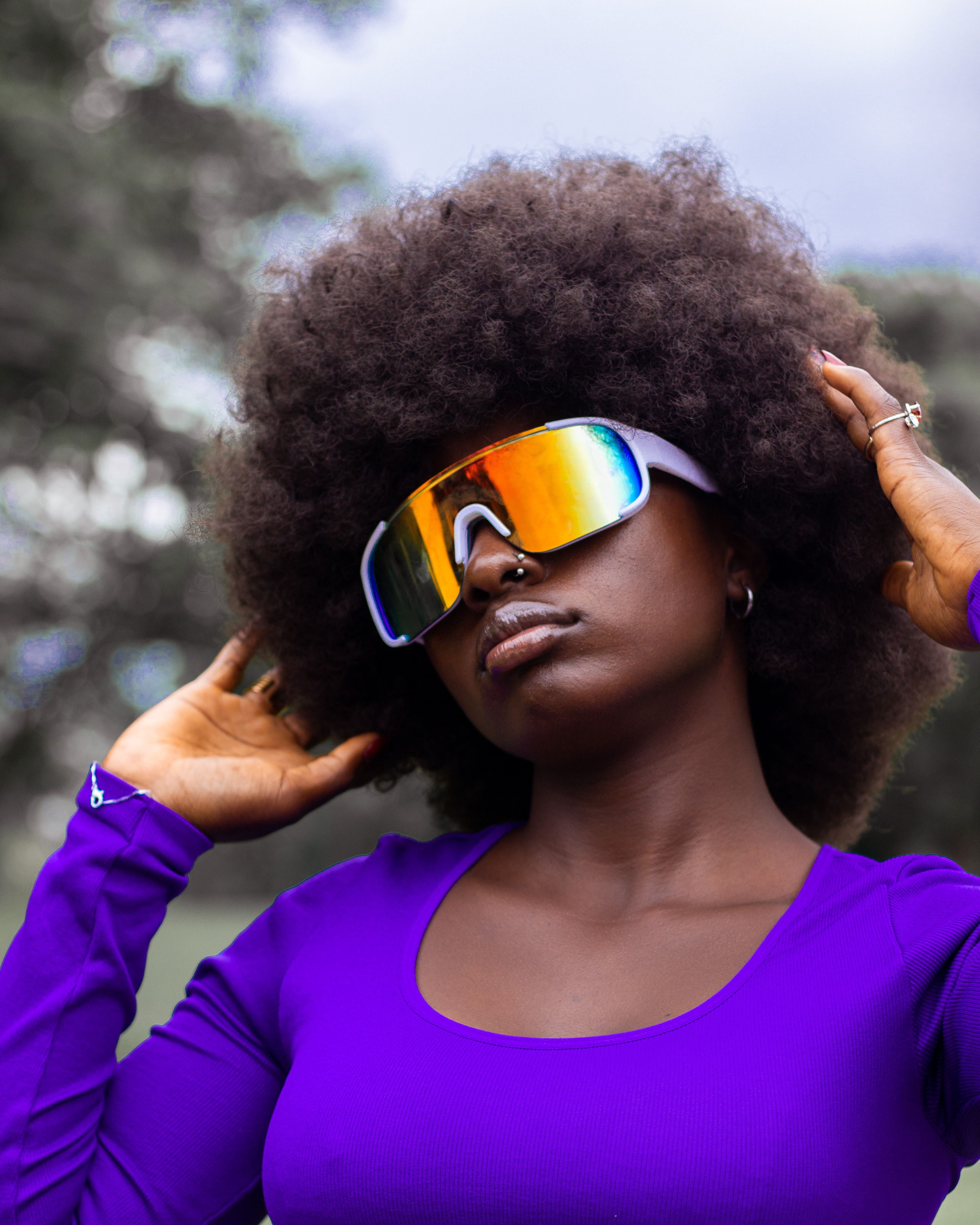 A fashionable woman with afro hair and futuristic sunglasses poses confidently outdoors.