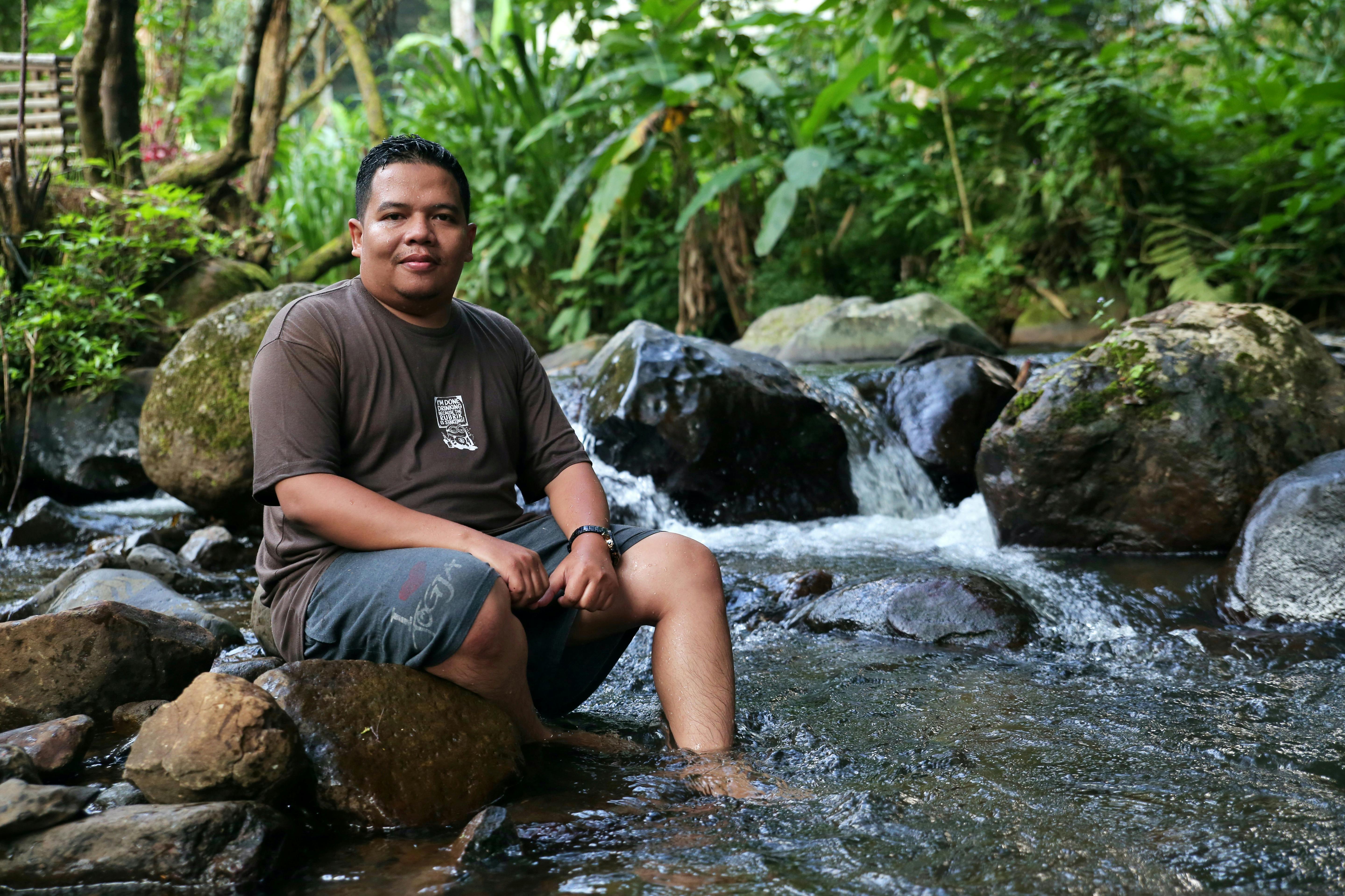 Adult man sitting in a natural stream surrounded by lush greenery and rocks.