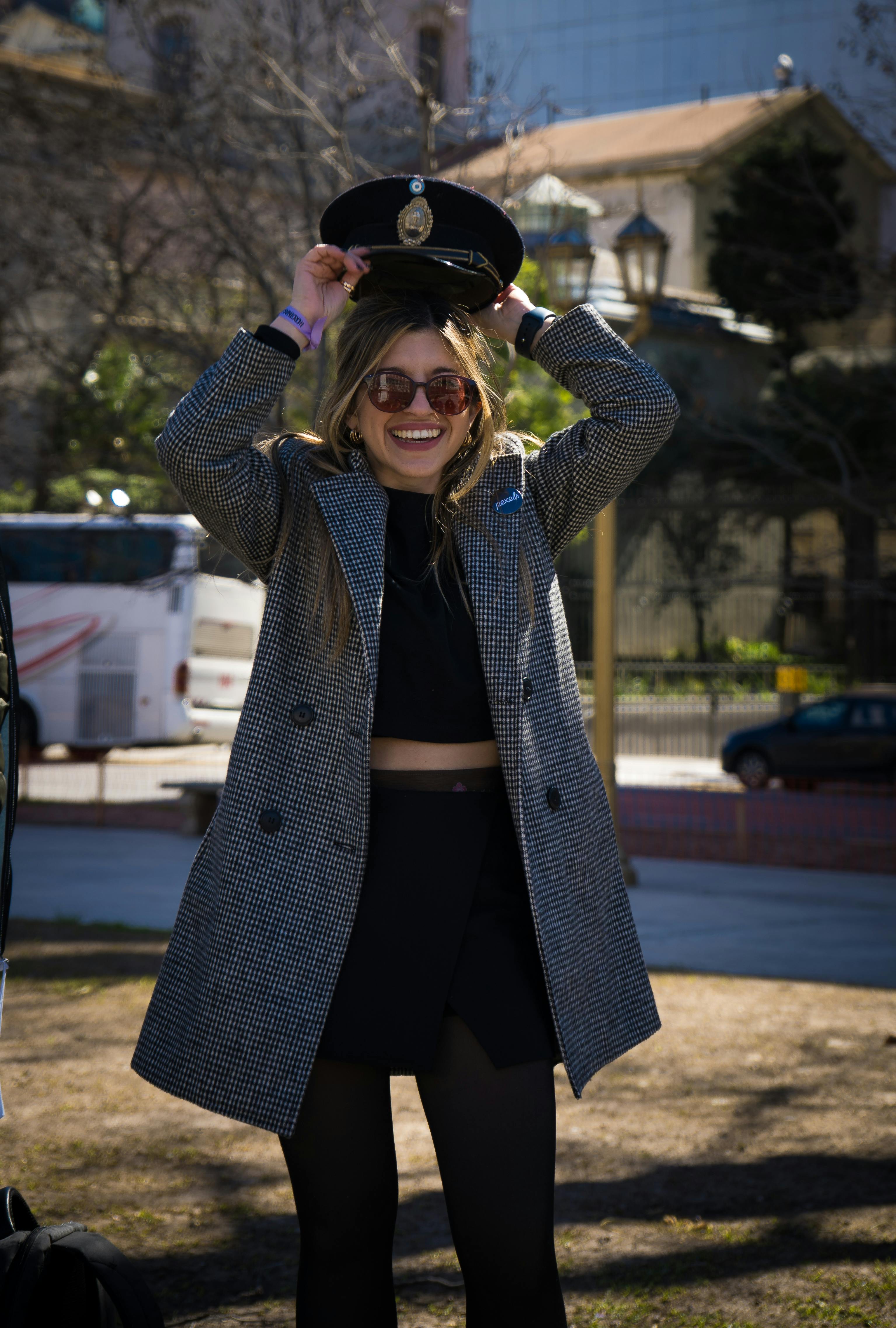 Smiling young woman in stylish coat holding hat outdoors on a sunny day.