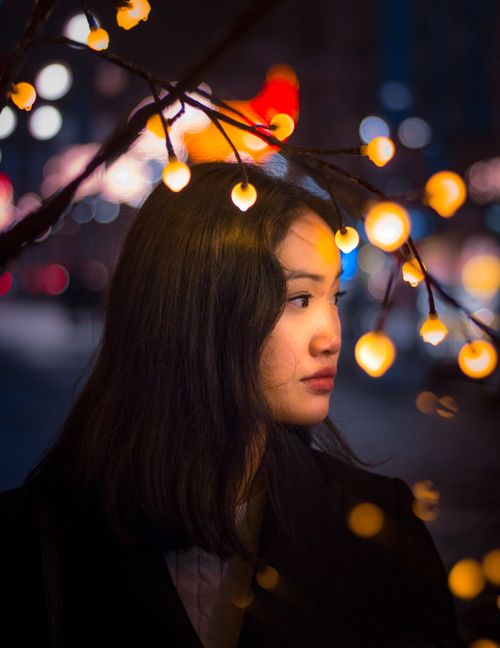 Free Asian woman gazes thoughtfully with city street and festive lights in the background. Stock Photo