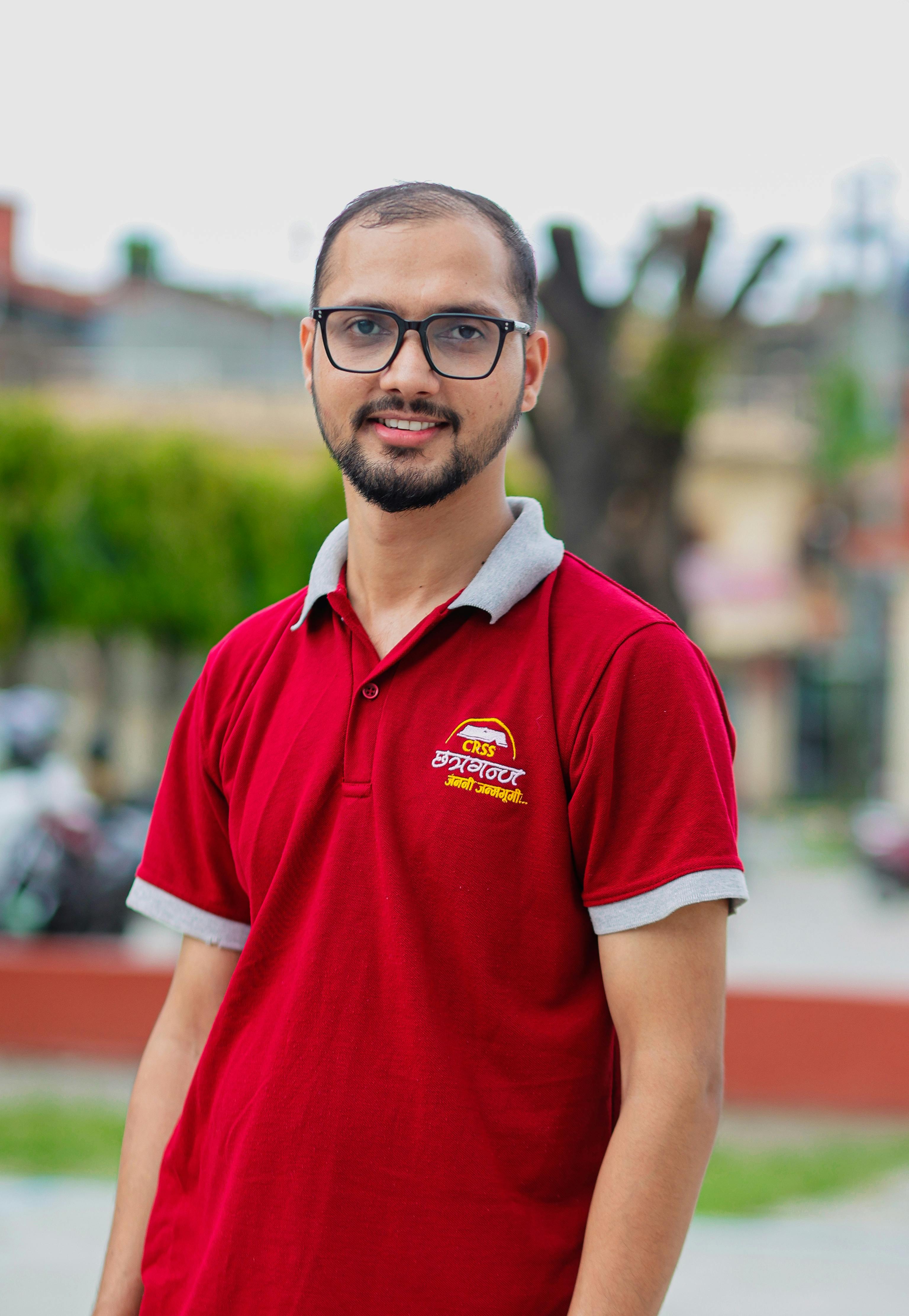 Smiling man in red shirt poses casually outdoors. Bright and cheerful setting.