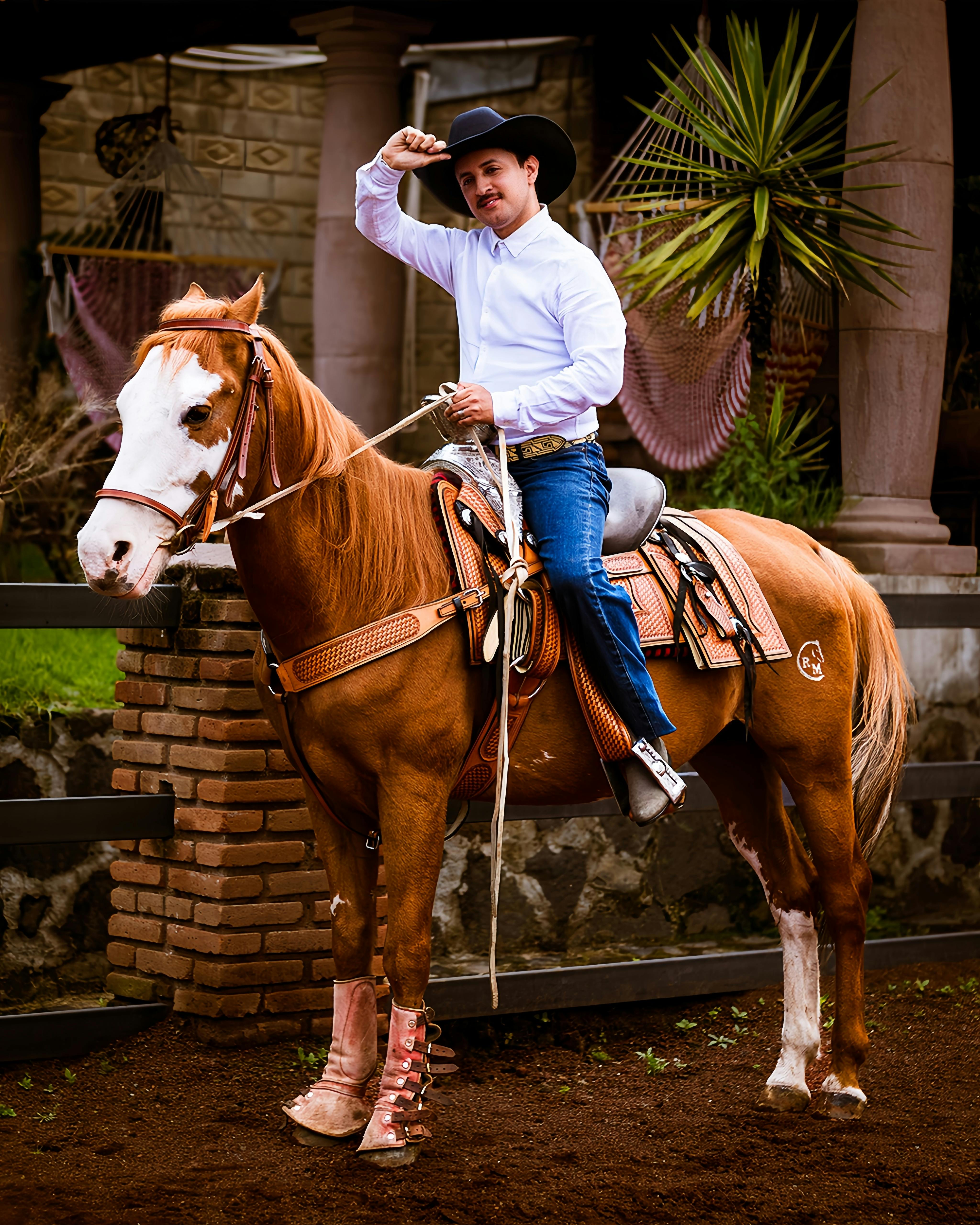 A cowboy in white shirt and hat rides a brown horse at a ranch, exuding western charm.