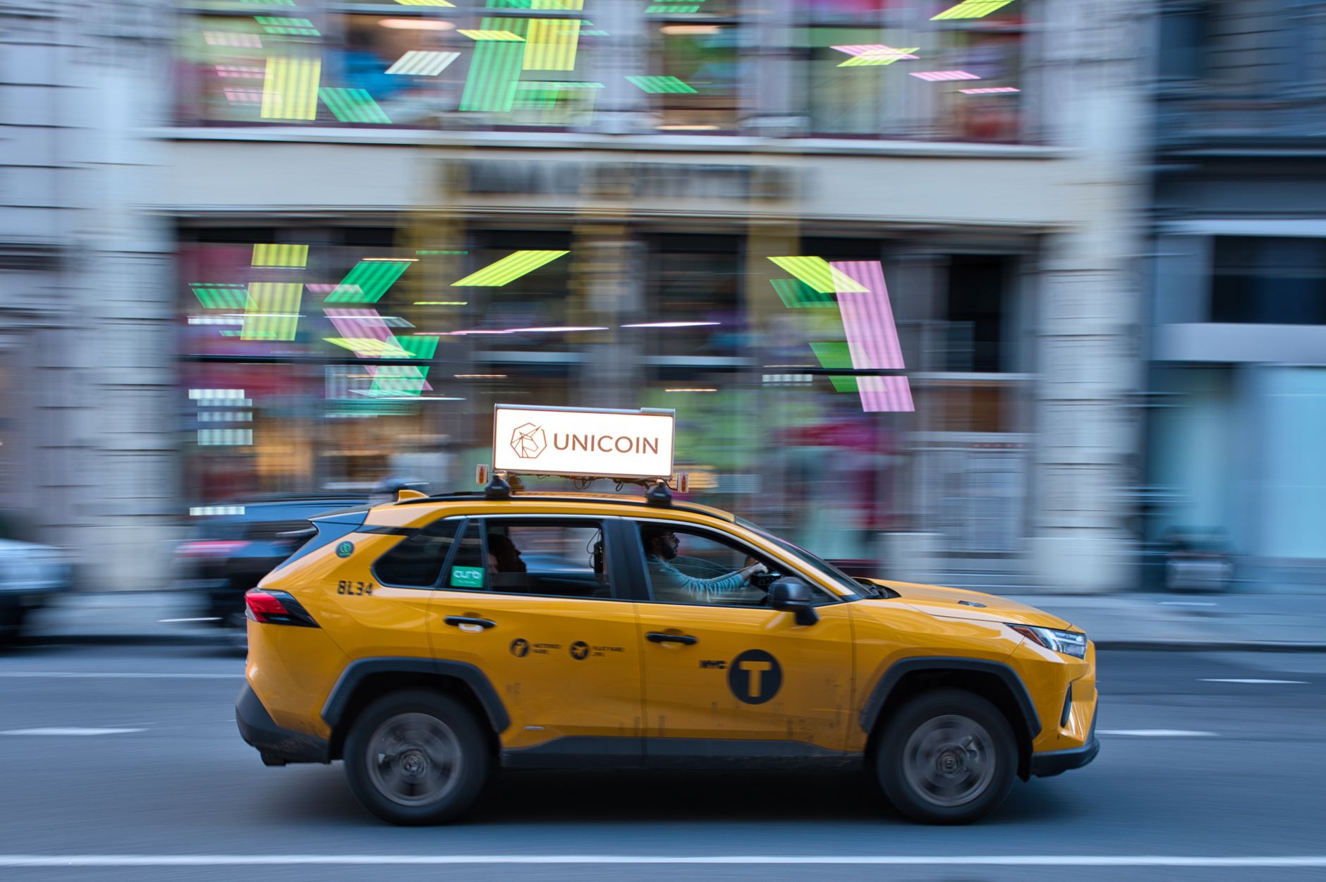 Yellow taxi in New York City speeds past a colorful building at dusk.