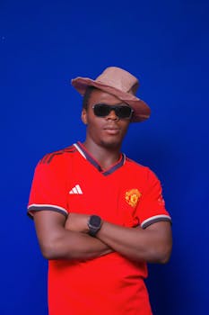Man wearing a red sports jersey, hat, and sunglasses against a blue background.