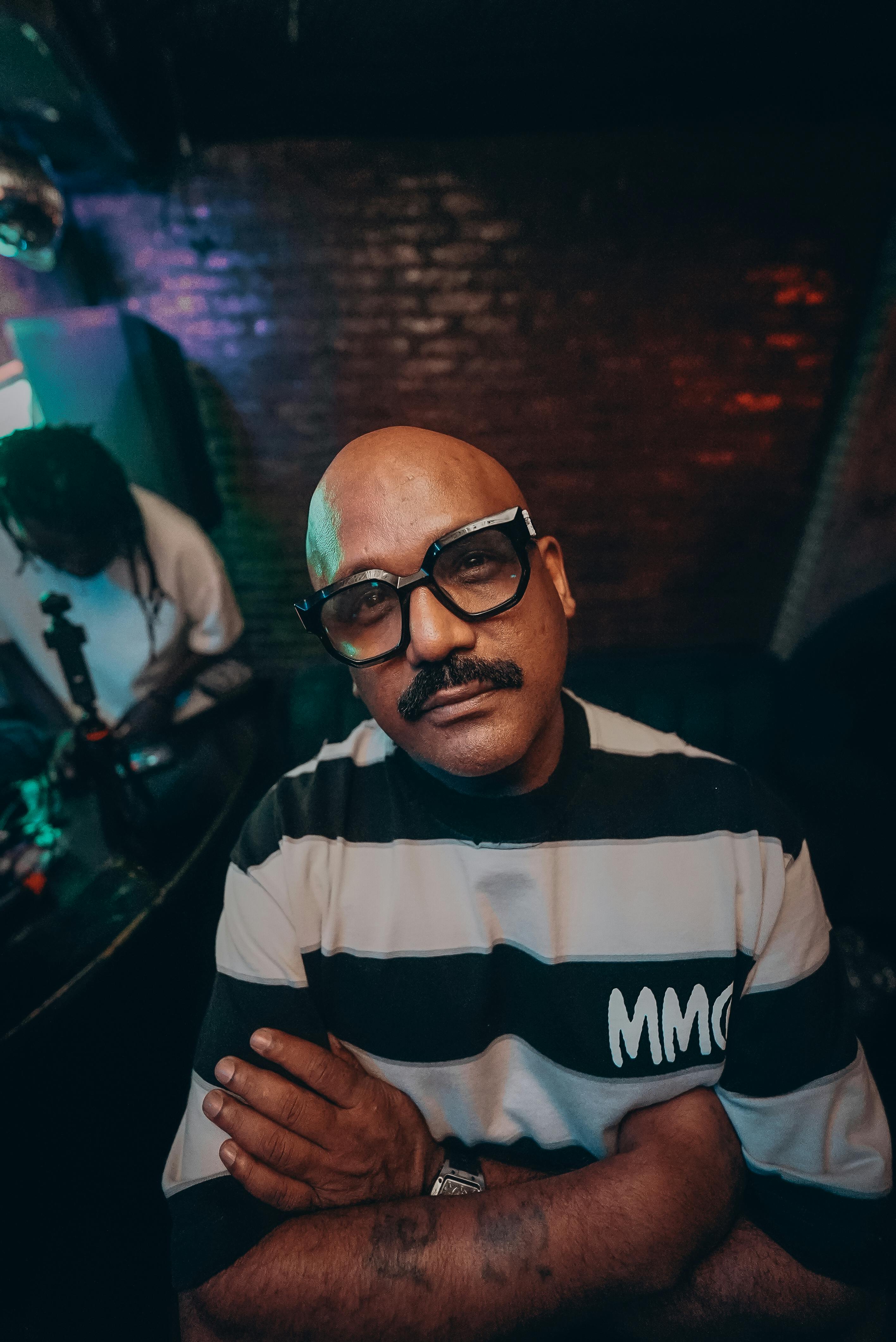 Confident man with glasses poses indoors in stylish striped shirt under colorful lights.