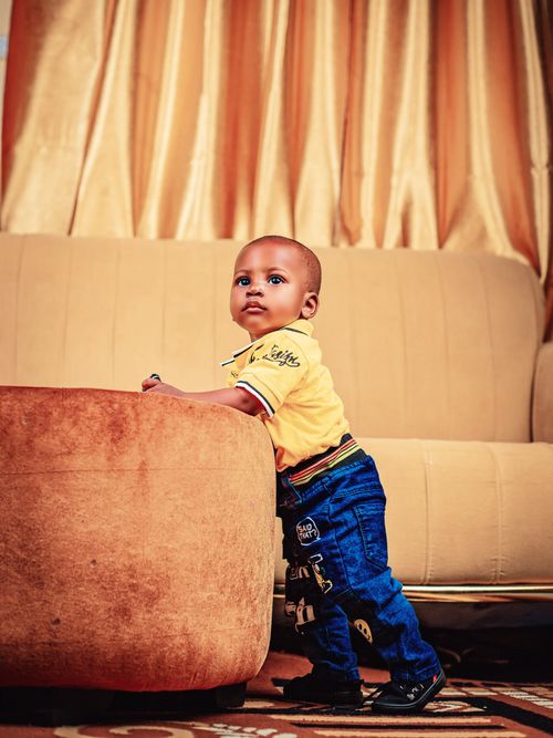 Free Cute baby boy in yellow shirt and jeans stands by a couch in a warm-toned living room. Stock Photo