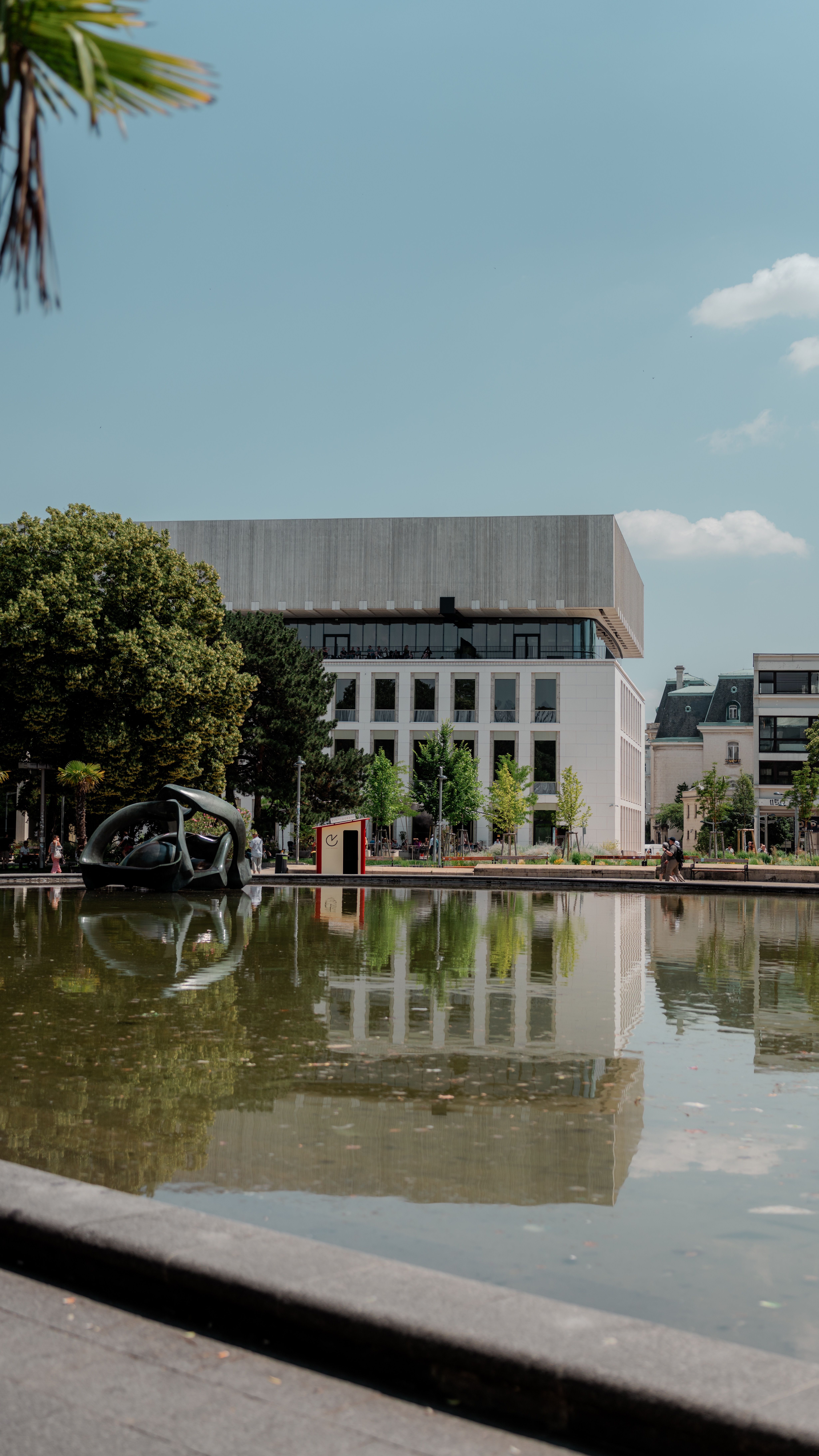 Contemporary building reflected in a pond on a sunny day in an urban park setting.