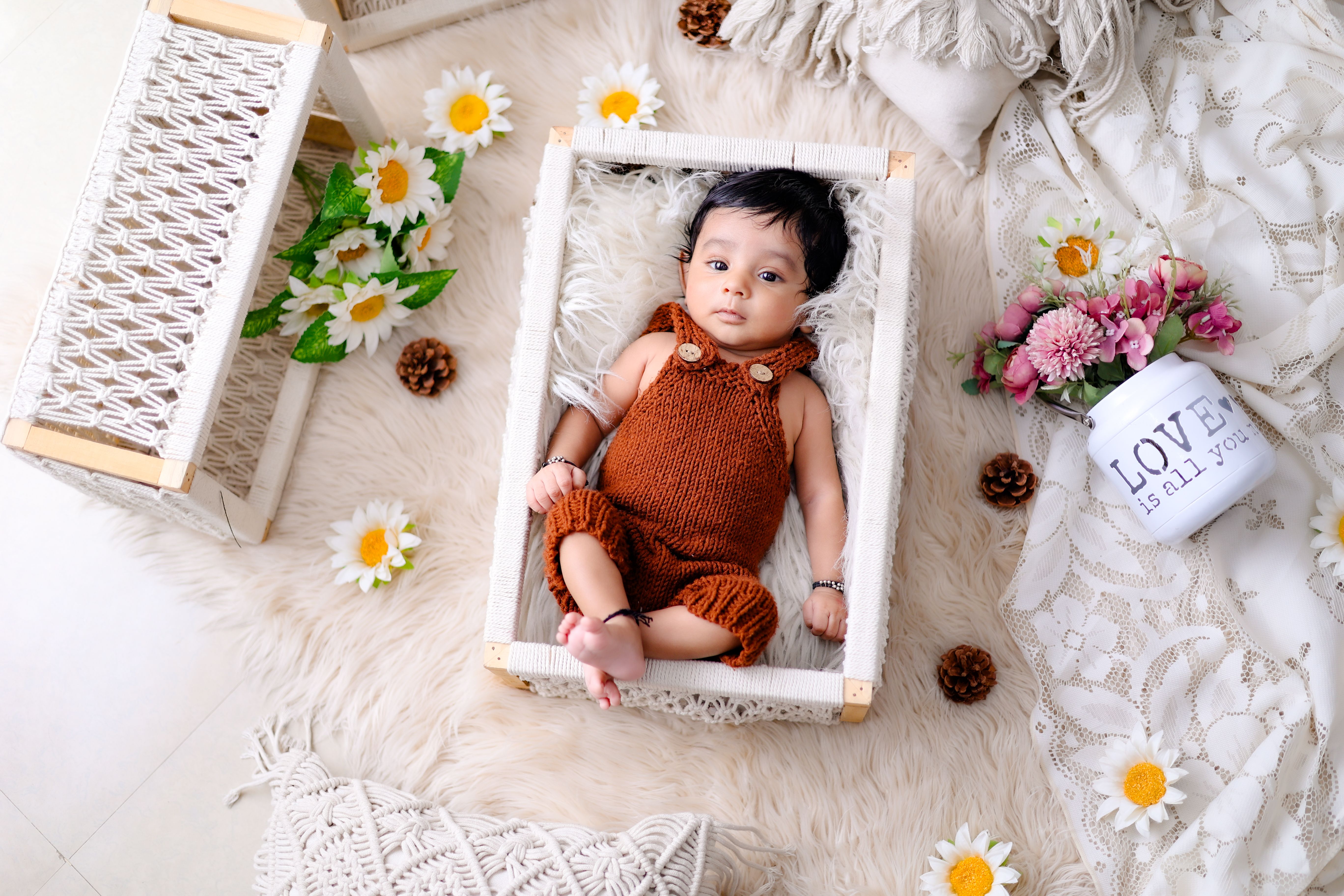 Charming baby in a knitted outfit surrounded by flowers and decor in a cozy indoor setting.