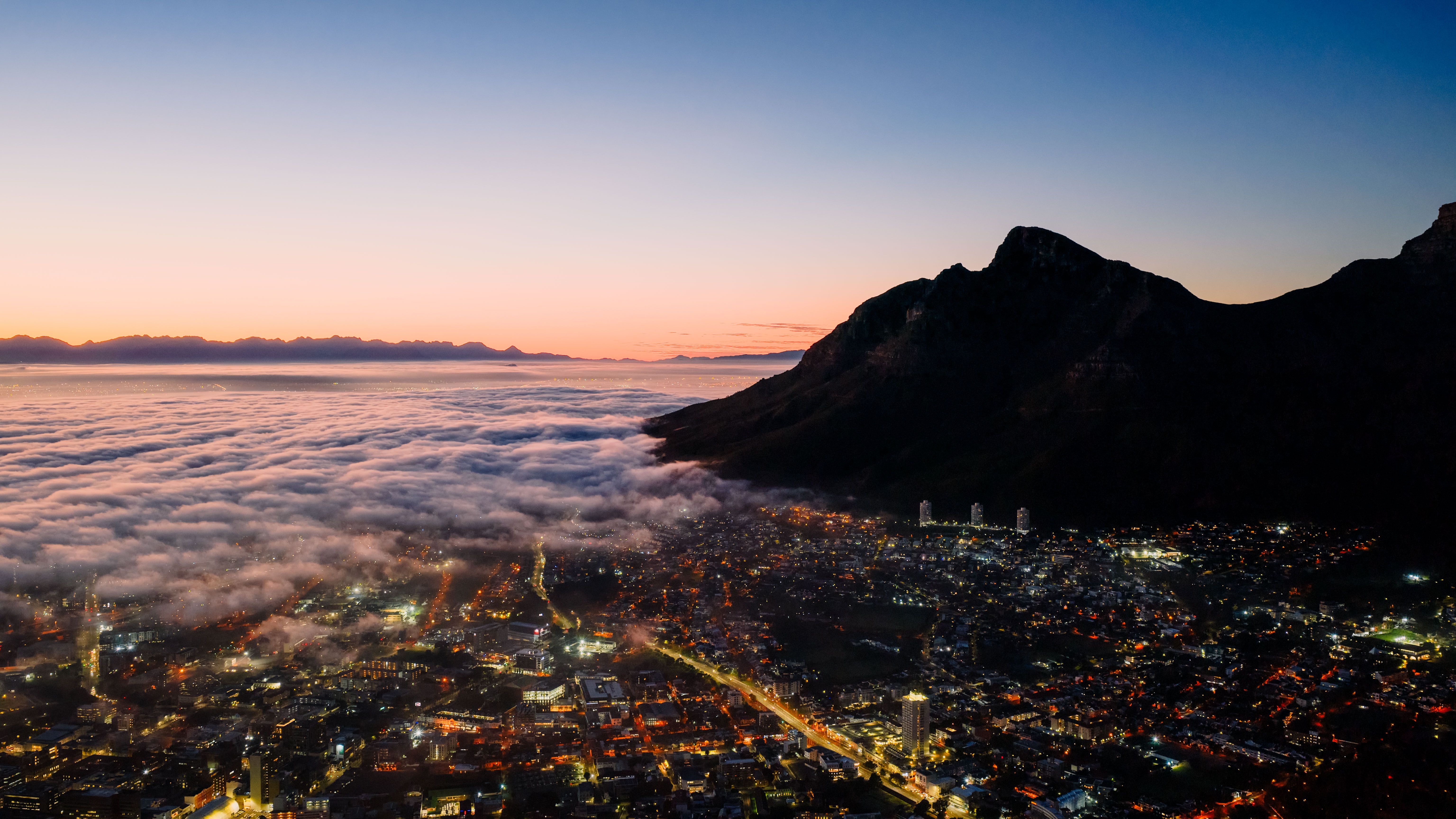 Unbeatable Cape Town City Center Tour with Champagne Review Review 2026 - Stunning aerial view of Cape Town with cloud cover and mountains at twilight, showcasing vibrant city lights.