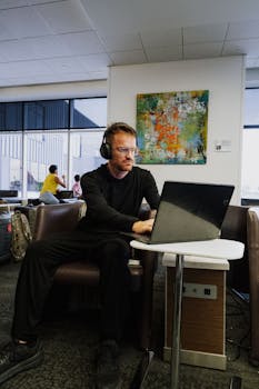 Business traveler wearing headphones works on laptop in airport lounge, focusing on productivity.