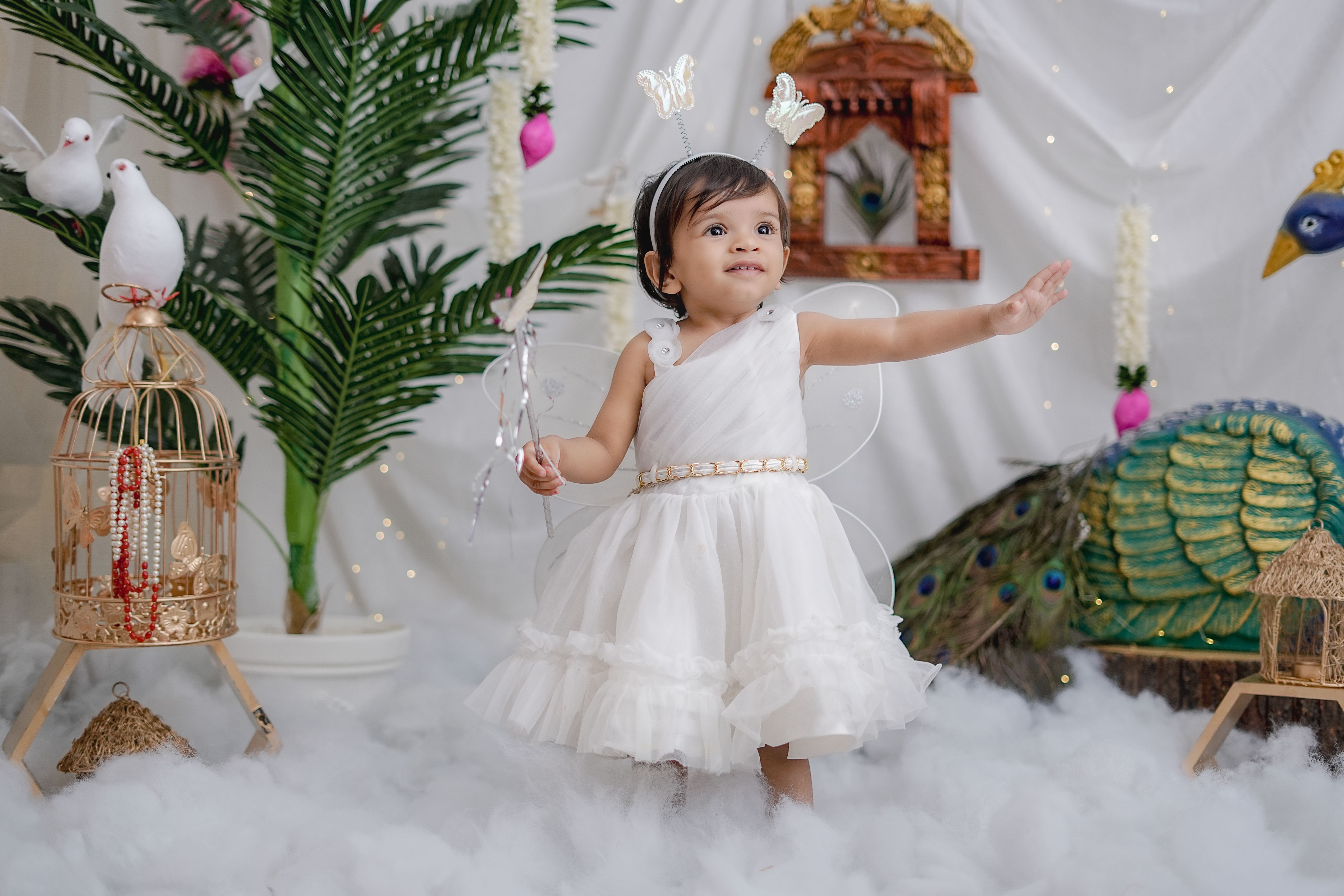 Charming child dressed as a fairy with whimsical background, including peacock and bird decor, creating a magical scene.