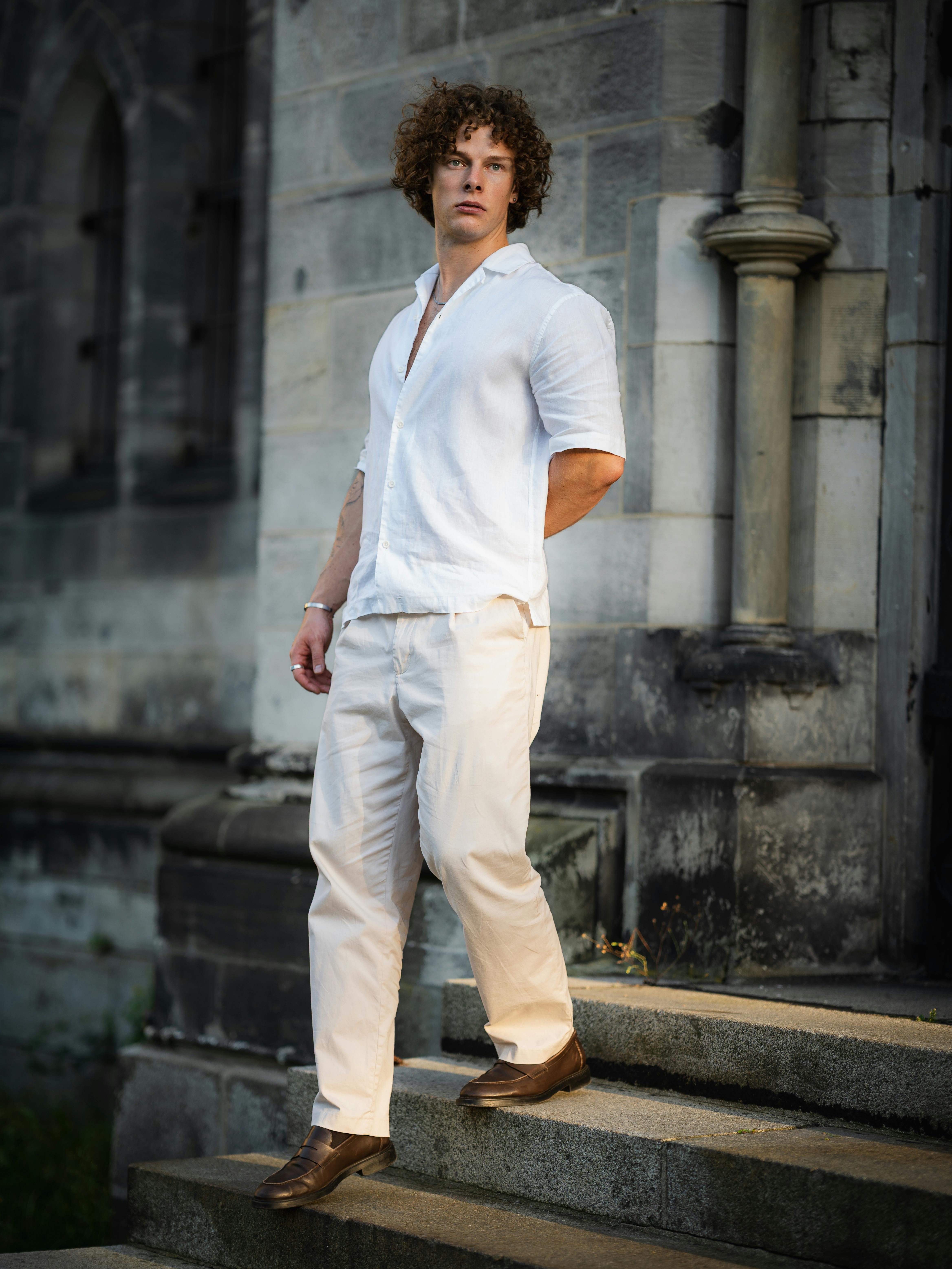 A fashionable man in light attire poses confidently on stone steps by a historic building.