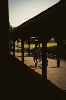 A person walking with a bicycle at Zittau train station, casting long shadows in the summer light.