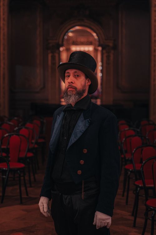 Free A man in historical costume with a top hat stands in a dimly lit ornate hall. Stock Photo