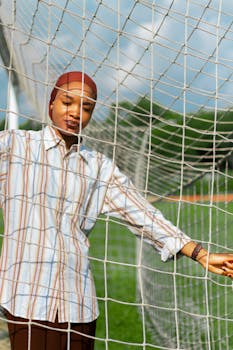 A young woman in a hijab stands by a soccer net outdoors, enjoying a sunny day.