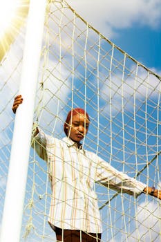 Young adult standing by a soccer net in sunlight against blue sky.