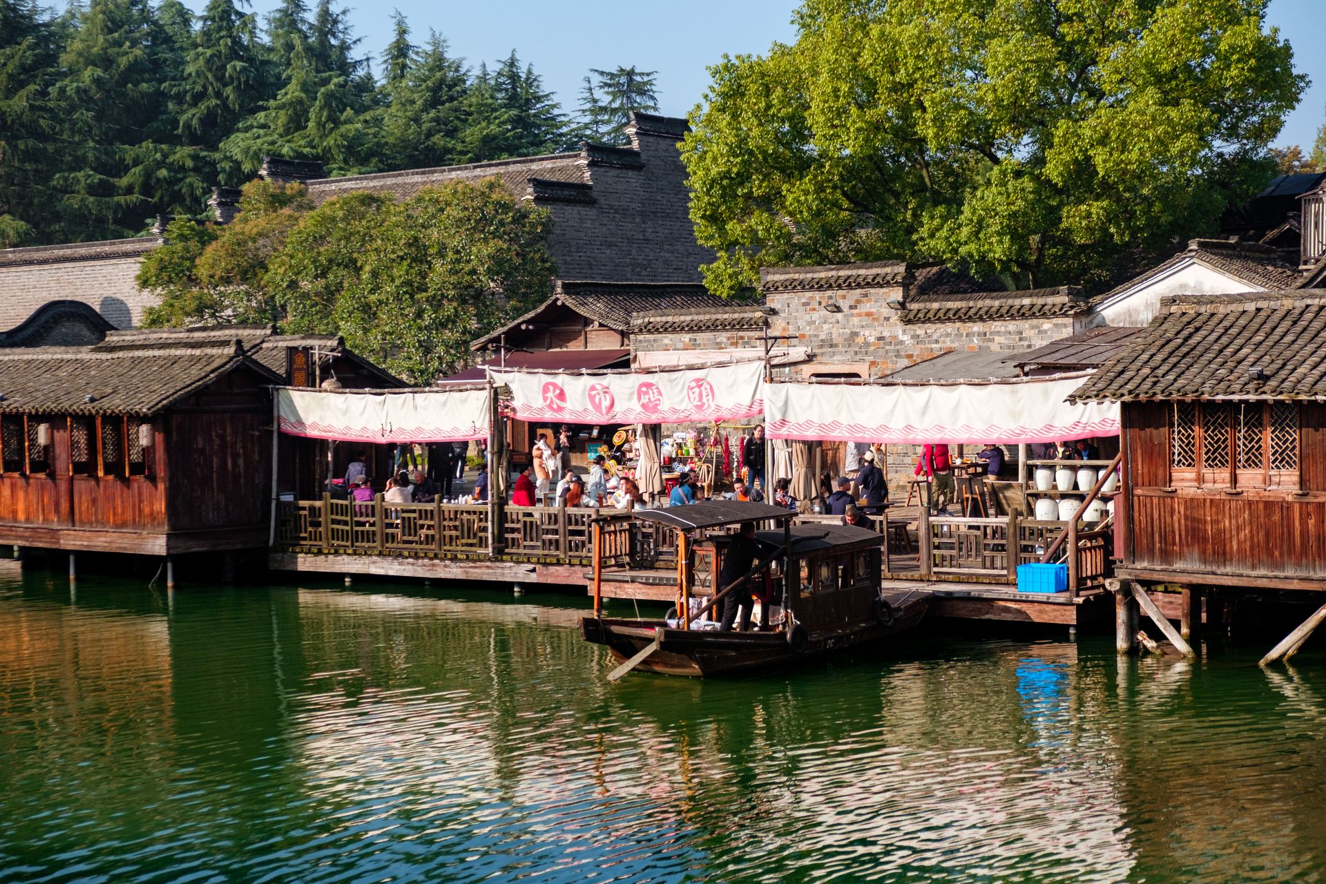 Charming riverside market with wooden structures and lush greenery in an Asian village setting.